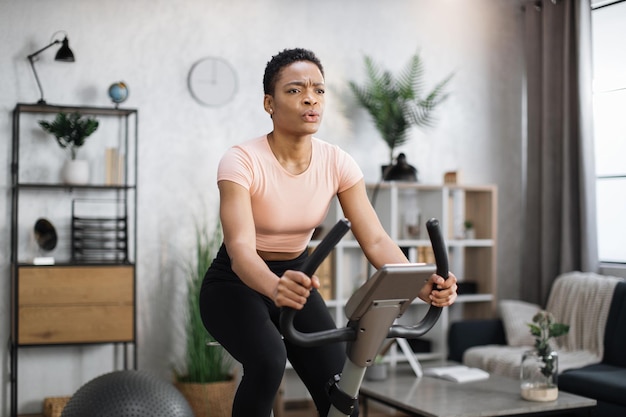 Retrato de una joven atleta africana enfocada que usa ropa deportiva y usa bicicleta de ejercicio