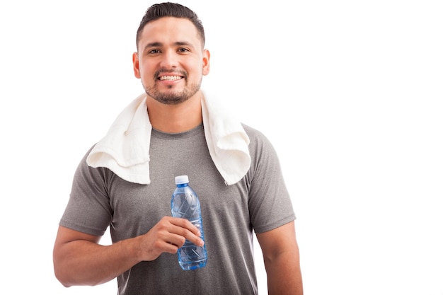 Retrato de un joven atleta hispano con una toalla alrededor del cuello y una botella de agua relajándose y refrescándose en un fondo blanco