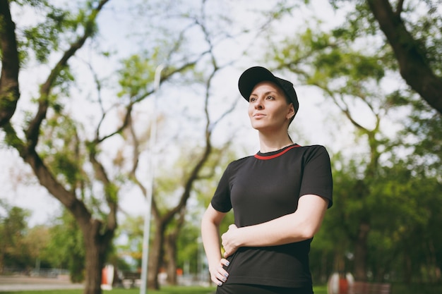 Retrato de joven atlética hermosa mujer morena en uniforme negro y gorra de pie, descansando y mirando a un lado después de correr, entrenando en el parque de la ciudad al aire libre