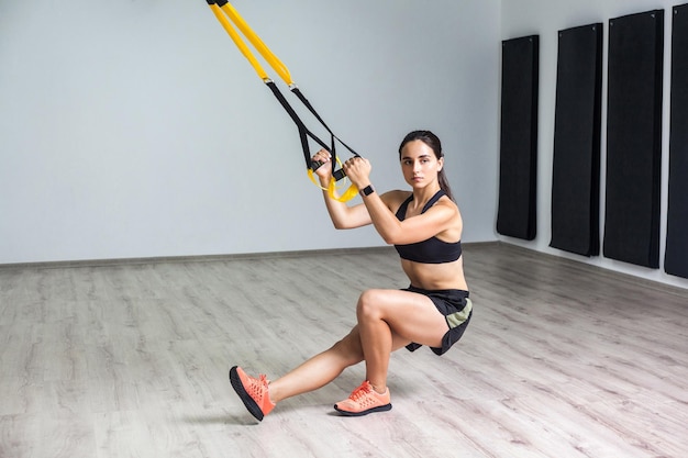 Retrato de joven atlética en ropa deportiva haciendo ejercicio en cuclillas entrenando piernas y glúteos musculares con correas de fitness en el gimnasio Entrenamiento funcional Concepto trx aislado fondo de pared blanca