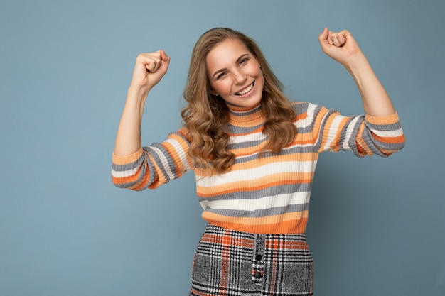 Retrato de joven atractiva atractiva feliz sonriente mujer rubia con cabello ondulado vistiendo suéter a rayas aislado sobre fondo azul con espacio vacío y celebrando la victoria. Concepto de victoria.