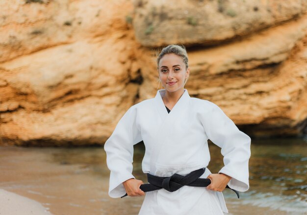 Retrato de joven y atractiva mujer maestra de artes marciales en kimono blanco con cinturón negro en la playa salvaje