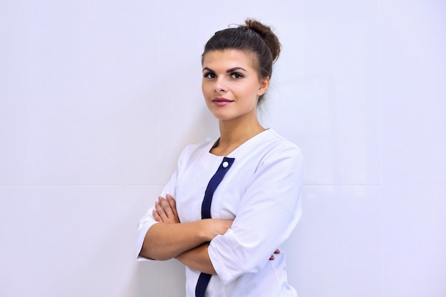 Retrato de joven atractiva mujer segura en uniforme médico con los brazos cruzados, fondo de clínica de pared clara. Farmacéutica, enfermera, cosmetóloga, especialista, científica