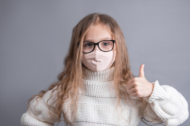 Retrato de una joven y atractiva niña con un suéter blanco en máscara protectora médica muestra los pulgares para arriba