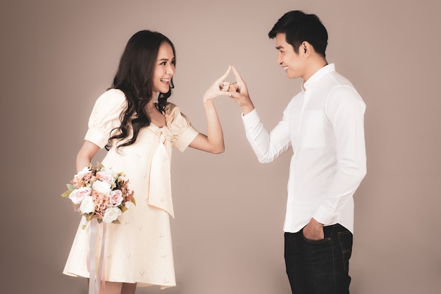 Retrato de joven atractiva pareja asiática, hombre vestido con camisa blanca, mujer con vestido beige de pie juntos jugando, mujer sosteniendo ramo de flores. Concepto de fotografía previa a la boda.