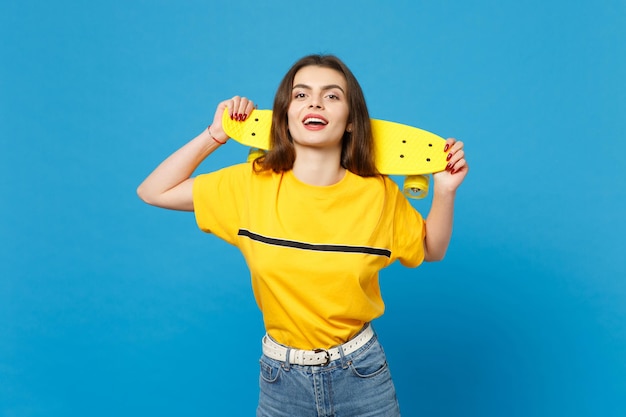 Retrato de una joven atractiva con ropa casual vívida de pie, mirando la cámara, sosteniendo una patineta amarilla aislada en el fondo de la pared azul en el estudio. Concepto de estilo de vida de las personas. Simulacros de espacio de copia.