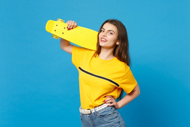 Retrato de una joven atractiva con ropa casual vívida de pie, mirando la cámara, sosteniendo una patineta amarilla aislada en el fondo de la pared azul en el estudio. Concepto de estilo de vida de las personas. Simulacros de espacio de copia.