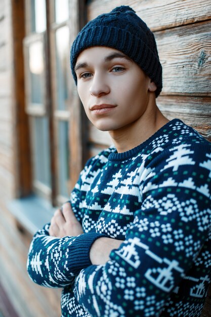 Retrato de un joven atractivo con hermosos ojos azules con piel bronceada en un sombrero azul de invierno con un suéter de punto con patrón de Navidad blanco cerca de una pared de madera vintage. Chico guapo.