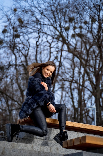 retrato, de, joven, atractivo, mujer, sentado, en, banco, en, parque, posar, bastante, niña, sonriente, en, invierno, en el parque, abrigo de piel oscuro, y, pelo largo