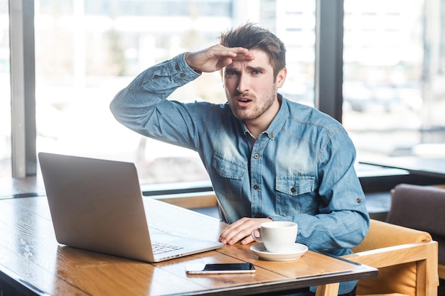 El retrato del joven autónomo barbudo hermoso se preguntó en la camisa de los pantalones vaqueros azules está sentado en la cafetería y trabajando en la computadora portátil, sosteniendo la mano cerca de la frente con cara de sorpresa, mirando a la cámara. interior