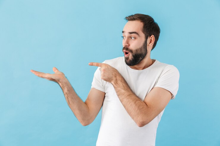 Foto retrato de un joven barbudo conmocionado vestido con camiseta que se encuentran aisladas sobre la pared azul, presentando espacio de copia
