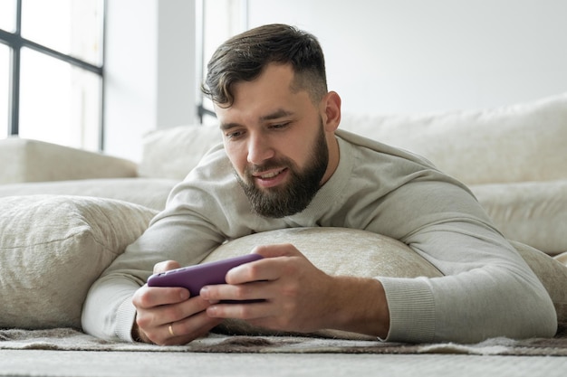 Retrato de un joven barbudo emocionado tirado en el suelo y jugando en un teléfono móvil