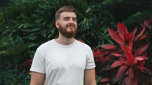 Retrato de un joven barbudo feliz con barba respirando profundamente inhalar aire fresco en la pared verde de fondo natural con flores de hojas en un país tropical en el jardín Concepto ecológico ecológico