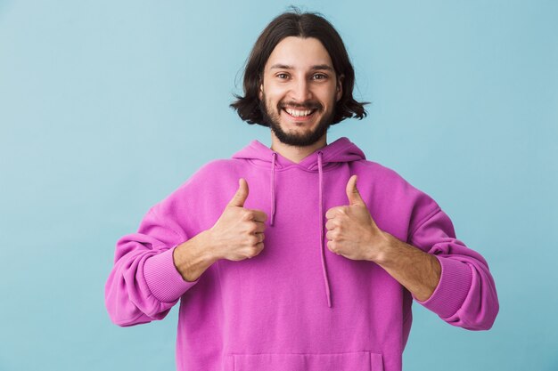 Retrato de un joven barbudo morena hombre vestido con sudadera con capucha que se encuentran aisladas sobre la pared azul, mostrando los pulgares para arriba