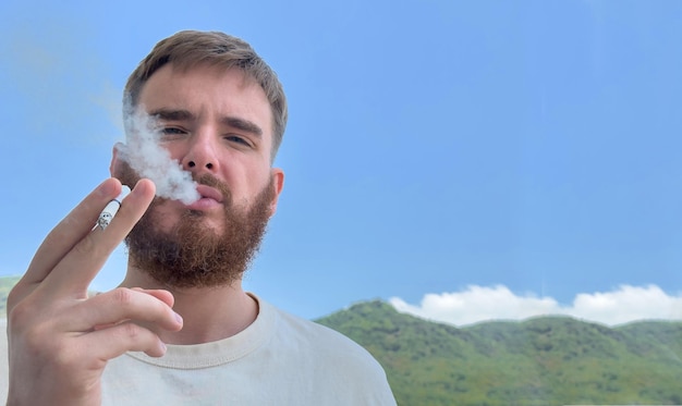 Retrato de un joven barbudo serio fumando cigarrillos en la terraza del balcón en el día de verano y mirando a la cámara Dejar de fumar concepto de mal hábito Fondo natural