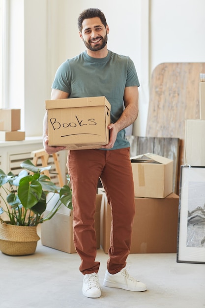 Foto retrato de joven barbudo sosteniendo una caja de cartón con libros y sonriendo mientras está de pie en su apartamento