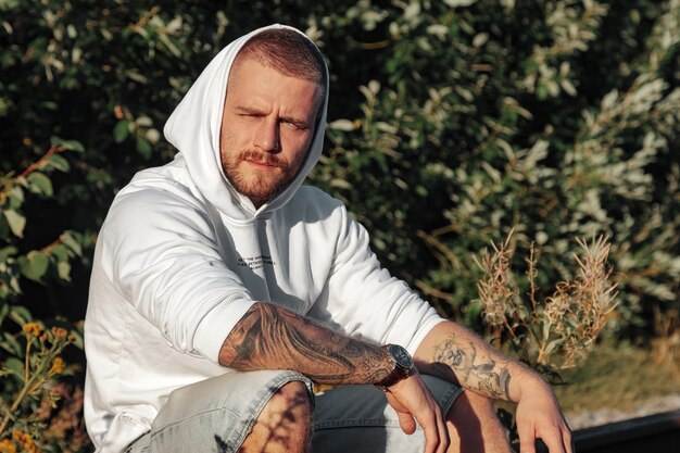 Retrato de joven barbudo con tatuajes en chaqueta blanca en campo o en el parque. Hombre en ropa casual pasa la noche en la naturaleza. Concepto de estilo, caminar al aire libre y unidad con la naturaleza.