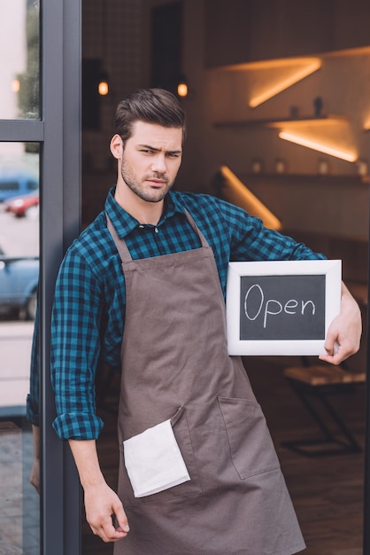 Retrato de joven barista en delantal sosteniendo pizarra con palabra abierta