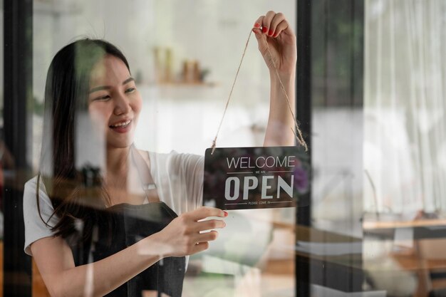 Foto retrato de una joven barista sonriente con un delantal sosteniendo un cartel abierto mientras está de pie en su cafetería