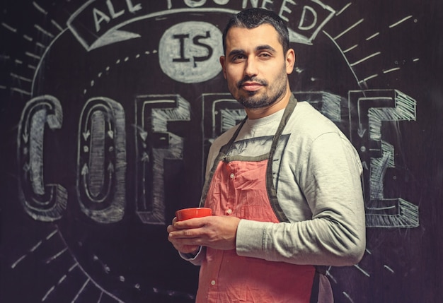 Retrato de un joven baristas en un delantal rojo con una taza en las manos de una pequeña cafetería.