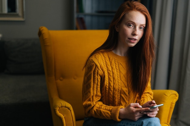 Retrato de una joven bastante pelirroja usando un teléfono inteligente sentado en un sillón amarillo confiado mirando a la cámara