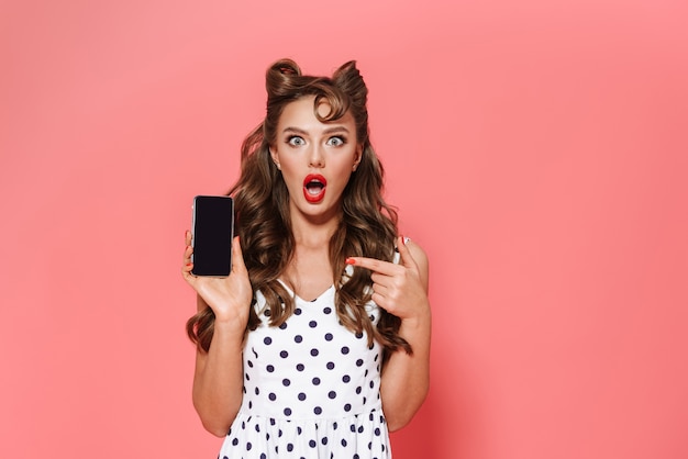 Retrato de una joven y bella chica pin-up con un vestido que se encuentran aisladas, mostrando el teléfono móvil de pantalla en blanco