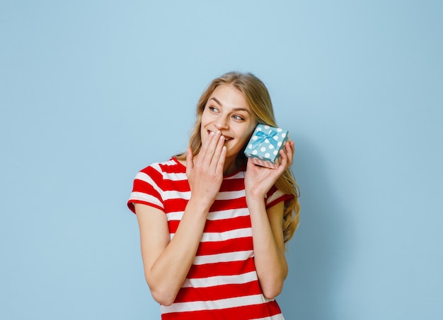 Retrato de una joven y bella chica rubia sosteniendo un regalo y feliz sobre fondo azul.