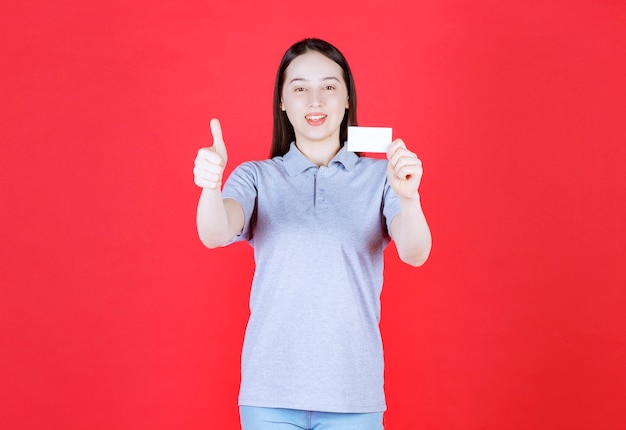 Retrato de joven bella dama sosteniendo la tarjeta de visita y gesticulando con el pulgar hacia arriba en la pared roja