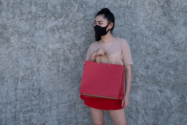 Retrato de una joven y bella mujer de Asia con una máscara facial con bolsas de compras en el muro de hormigón, las mujeres visten ropa rosa y falda roja