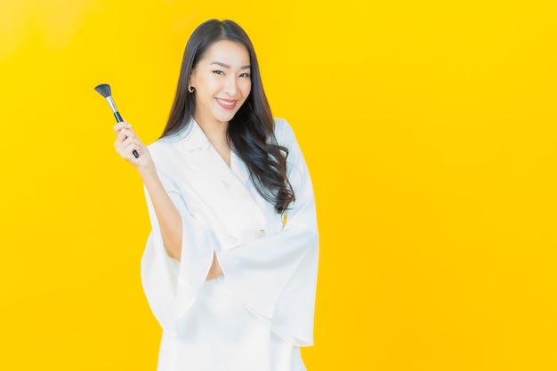 Retrato de joven y bella mujer asiática con maquillaje cosmético de pincel en pared amarilla