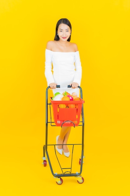 Retrato de joven y bella mujer asiática de negocios con cesta de la compra con comestibles del supermercado en la pared amarilla