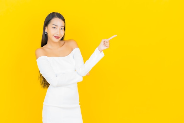 Retrato de joven y bella mujer asiática de negocios sonriendo y apuntando a un lado con traje blanco en la pared amarilla