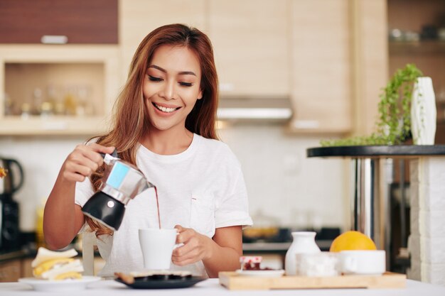 Retrato de joven y bella mujer asiática sirviéndose una taza de café cuando está de pie en la mesa de la cocina