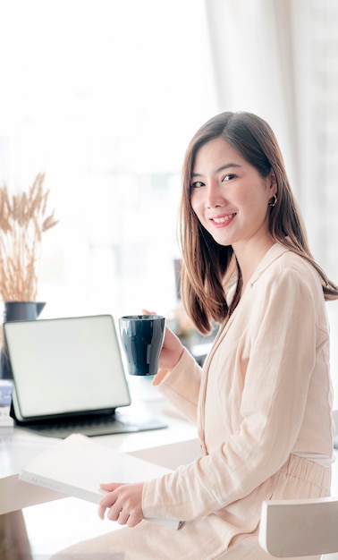 Retrato de joven bella mujer asiática sosteniendo la taza y el papeleo mientras está sentado en la oficina