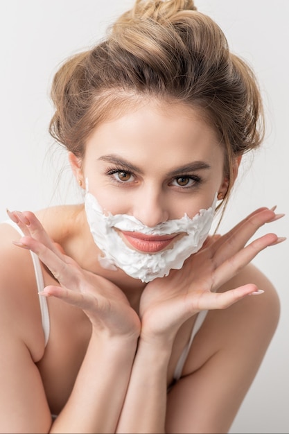 Retrato de una joven y bella mujer caucásica sonriente con espuma de afeitar en poses de cara sobre fondo blanco.