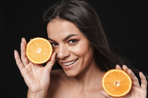 Retrato de una joven y bella mujer desnuda feliz posando aislada sobre pared negra con cítricos naranjas.