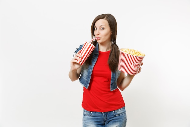 Foto retrato de joven bella mujer divertida en ropa casual viendo una película, sosteniendo un cubo de palomitas de maíz, bebiendo de un vaso de plástico de refresco o cola aislado sobre fondo blanco. emociones en el cine.