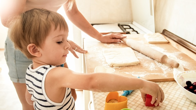 Retrato de joven bella mujer enseñando a su pequeño niño haciendo galletas y horneando pasteles en la cocina de casa