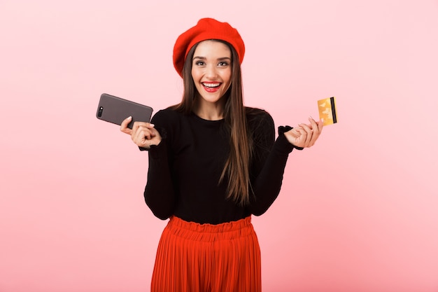 Retrato de una joven y bella mujer feliz vistiendo boina roja que se encuentran aisladas sobre fondo rosa, sosteniendo un teléfono móvil y una tarjeta de crédito de plástico