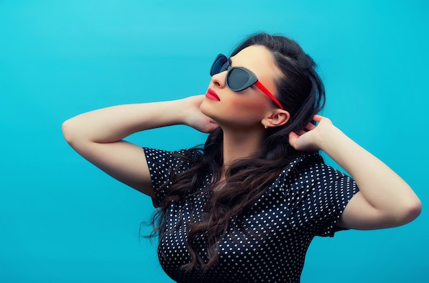 Retrato de la joven y bella mujer con gafas en el vestido de lunares negro en la pared azul. Modelo de mujer joven.