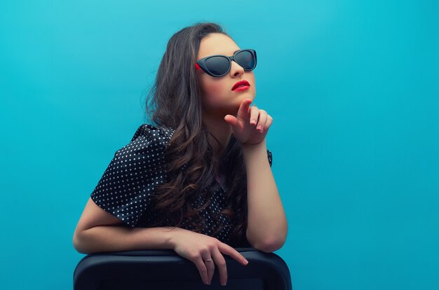 Retrato de la joven y bella mujer con gafas en el vestido negro de lunares vintage en la pared azul. Modelo de mujer joven.