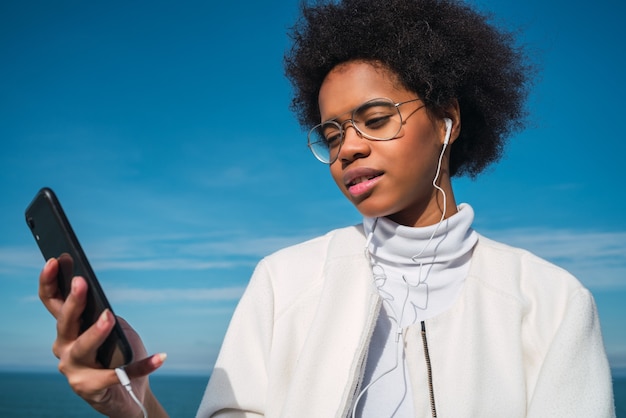 Retrato de joven bella mujer latina con su teléfono móvil con auriculares, con el mar en el espacio. Concepto de comunicación y tecnología.