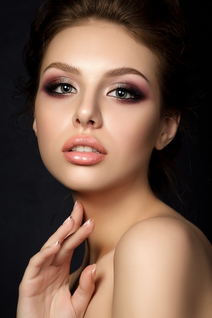 Retrato de joven bella mujer con maquillaje de noche tocando su rostro sobre fondo negro.