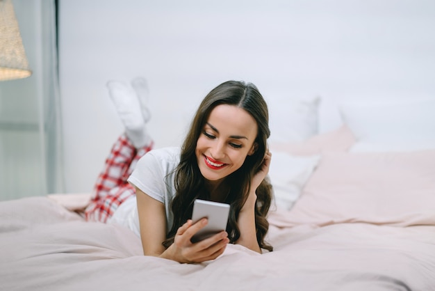 Retrato de una joven y bella mujer morena acostada en la cama navegando por su teléfono móvil