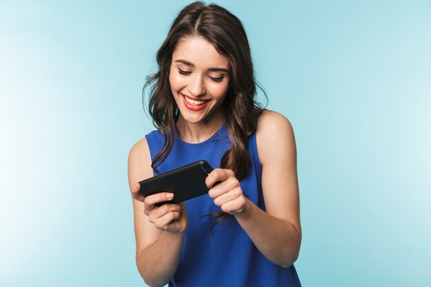 Retrato de una joven y bella mujer morena de pie sobre azul, jugando aplicaciones en el teléfono móvil