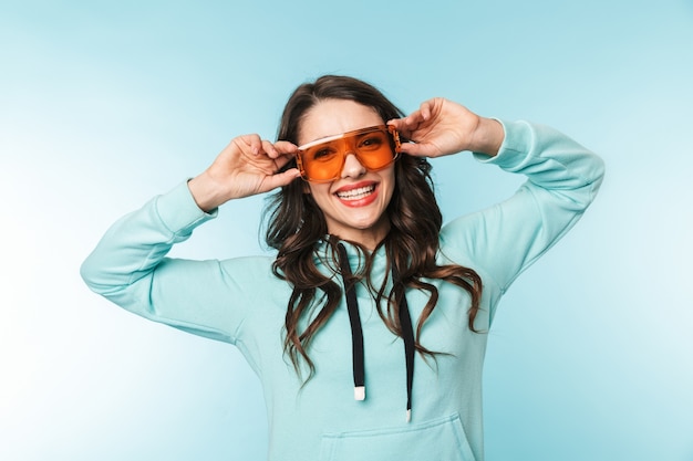 Retrato de una joven y bella mujer morena de pie sobre azul, posando con gafas de sol