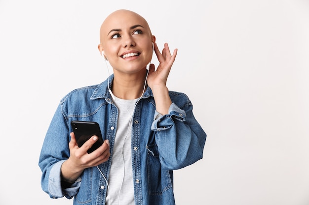 Retrato de una joven y bella mujer sin pelo con ropa casual que se encuentran aisladas, escuchando música con auriculares, sosteniendo el teléfono móvil