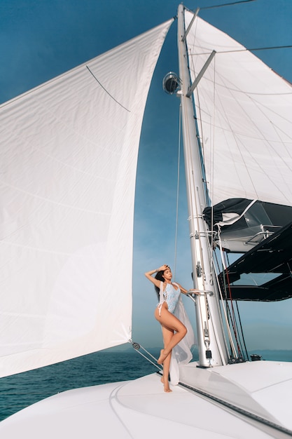 Retrato de joven bella mujer de pie y posando en velero o yate en el mar vistiendo traje de baño blanco moderno