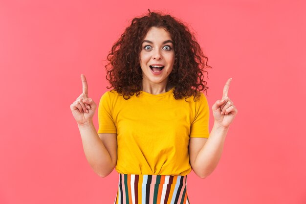 Retrato de una joven y bella mujer rizada posando aislada en la pared roja apuntando.