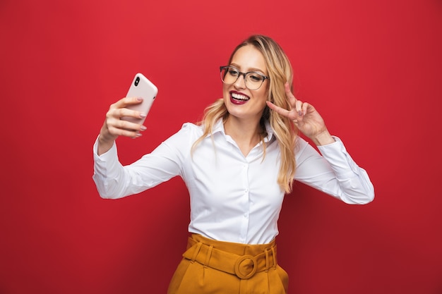 Retrato de una joven y bella mujer rubia que se encuentran aisladas sobre fondo rojo, tomando un selfie